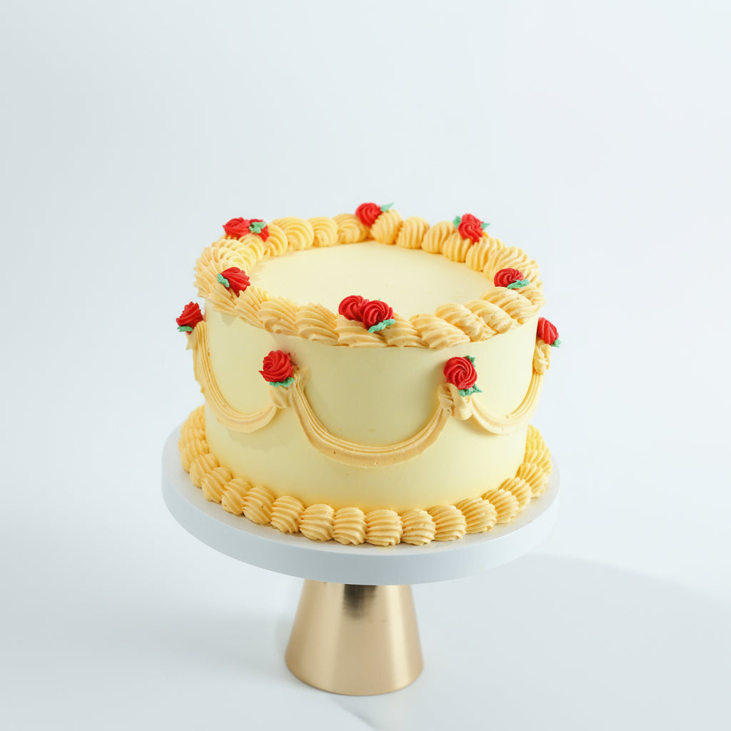 A two-tiered butter cream cake decorated with yellow icing and red rose decorations, presented on a cake stand.